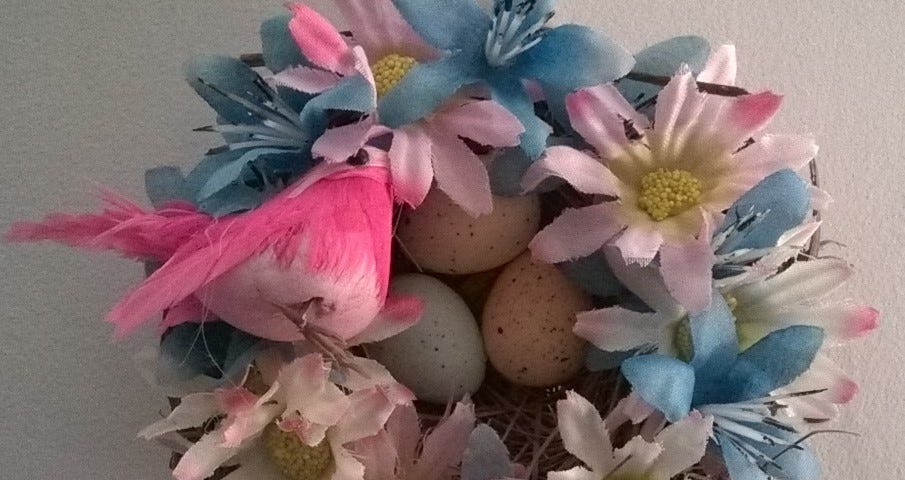 Pink and blue synthetic flowers cover a human-crafted bird nest filled with multicolored spotted eggs, while a pretty pink feathered bird sits to the side of the nest.