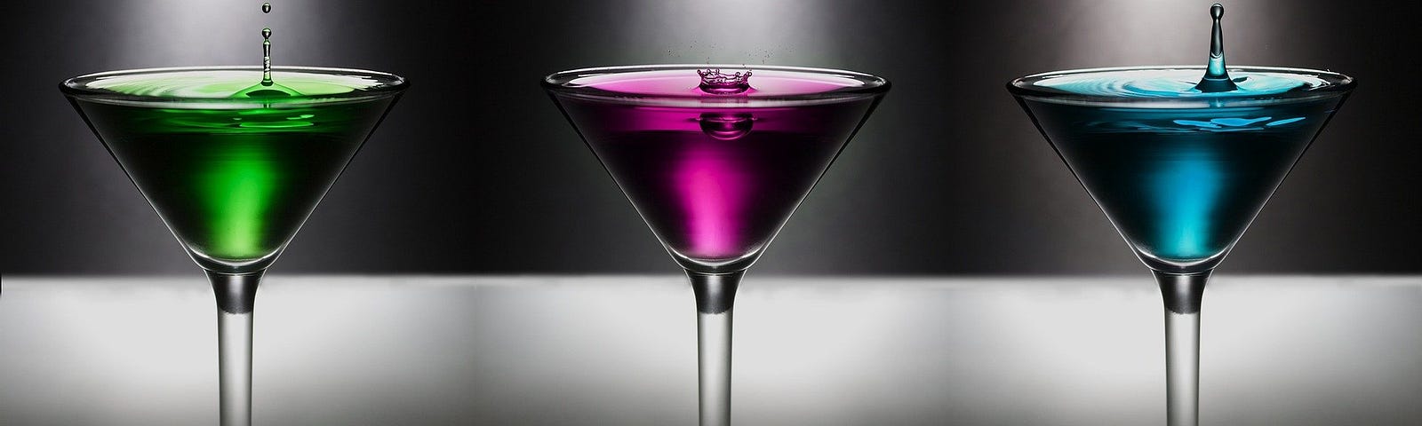 Three martini glasses side by side, filled with green, purple, and blue liquid respectively, on a gray background.