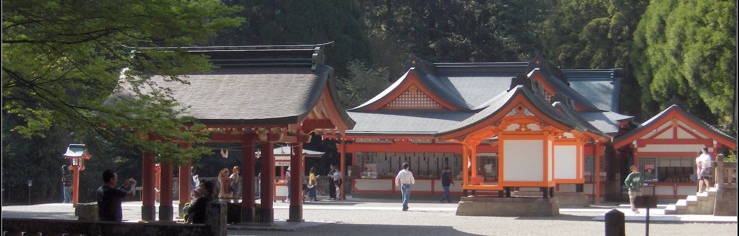 White shrine structures with vermilion accents and sloping rooflines amid a sunny courtyard with long shadows against a dense forested background.