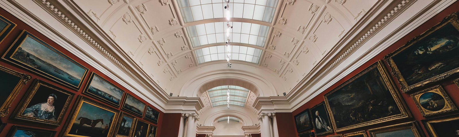 An art gallery room with high curved ceilings, parquet floor and old paintings in golden frames with a bench at the center to visitors to rest