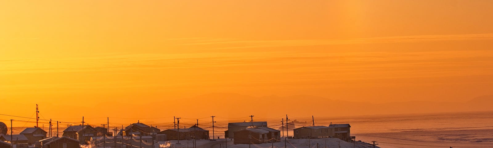 View of Pond Inlet Inuit community on Baffin Island. From a distance, orange and pink sky. Houses covered in snow.