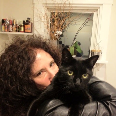 A woman with long, curly black hair wearing a black leather jacket (Karen Dendy Smith) and holding a black cat with yellow eyes (Hockey Puck).