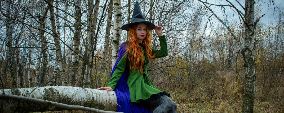 A young, attractive woman sits on a fallen tree trunk, among a thicket of silver birch. She is wearing a black ‘witch’ hat, a green minidress over grey leggings, a blue cape, and black ankle boots.