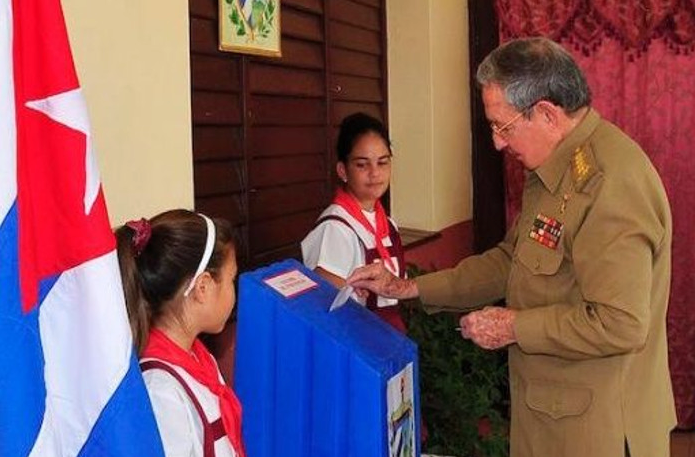 Former Cuban president Raúl Castro in military uniform casts a vote in a blue ballot box, flanked by two schoolchildren and the Cuban flag