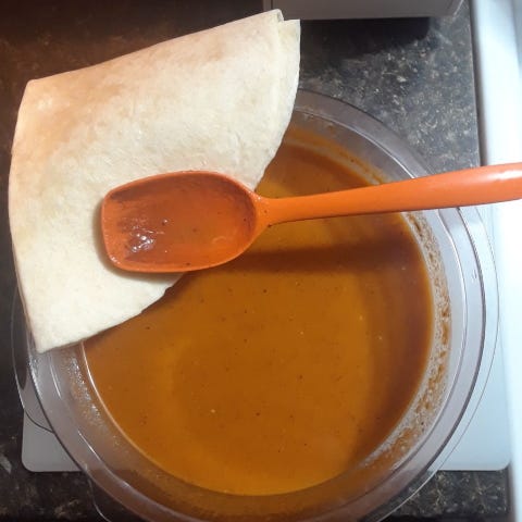 The final soup result, spices sparkling from the tomato mix, with the soup spoon sitting across the bowl, sitting partially on top a quartered soft shell.