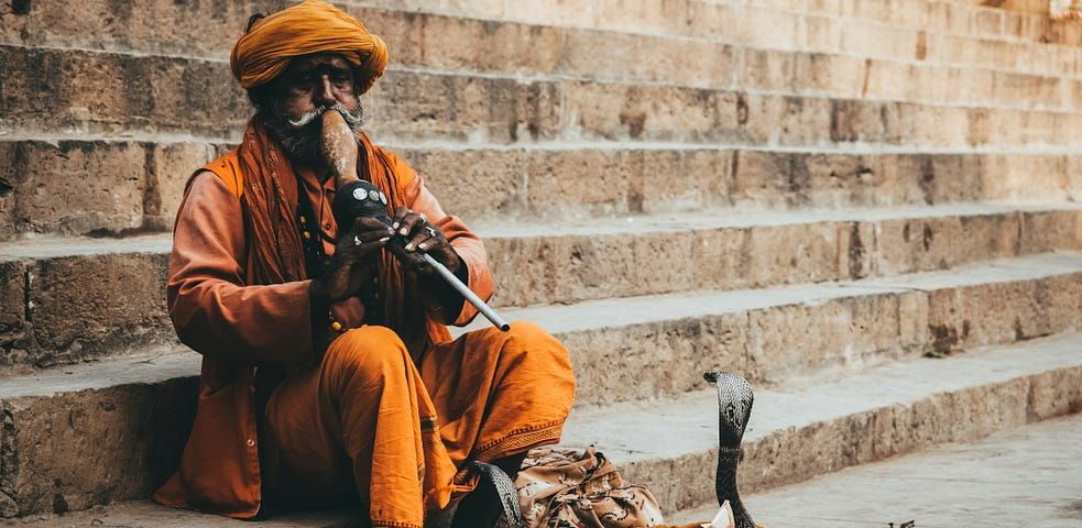 An Indian snake charmer in orange garb and turban, sitting on stone steps, playing his flute, mesmerizing a cobra, rising out of a basket.