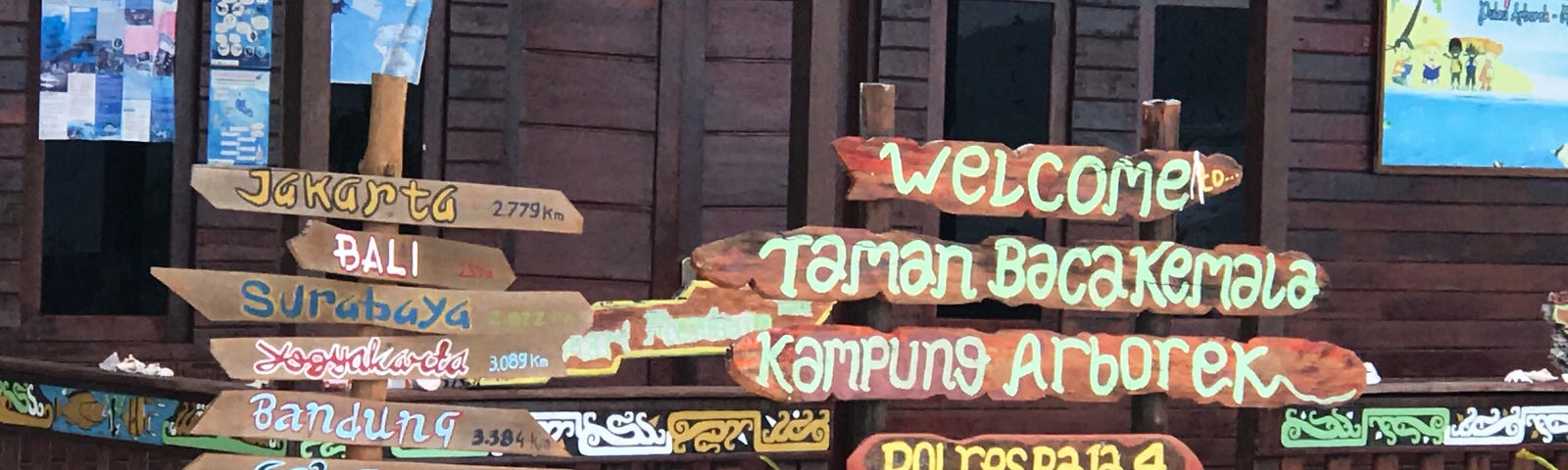 Wooden signs with directions pointing to distant travel spots in front of a wooden building. Signs say, “Welcome,” “Kampung Arborek,” “Bali,” etc.