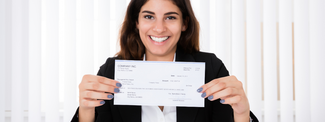 A woman with long brown hair with a part in the middle and a smile on her face. She is holding a paycheck with both hands. Her fingernail polish is grey-blue.