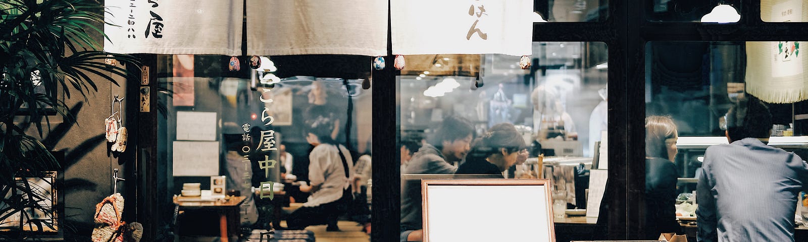 A photo of people dining at night in a Japanese restaurant. The photo is taken just outside of the restaurant, so the people eating inside can be seen.