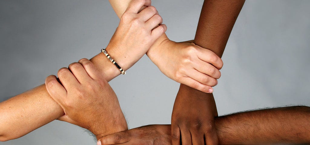 Five hands of people from varying races holding each other’s wrists forming a pentagon shape in the middle.