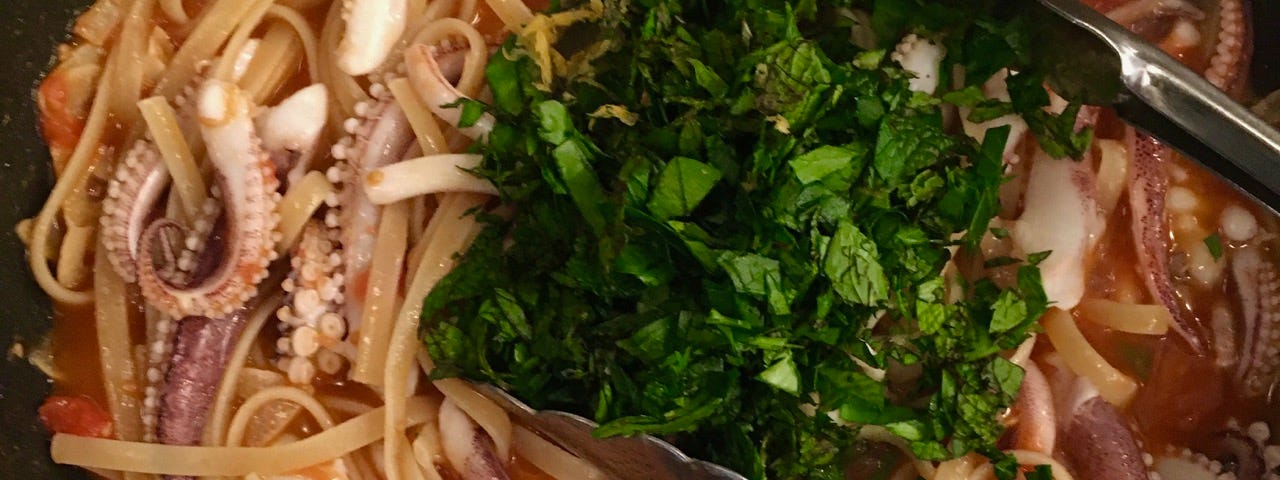 Overhead shot of a pan of linguini in sauce with squid tentacles on top, being topped with chopped herbs.