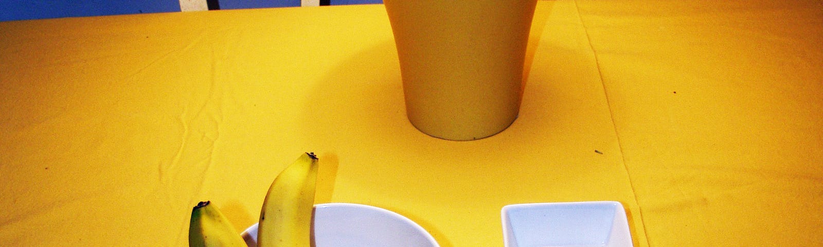 Dining room table with lamp, ceramic bowls and a couple of bananas. Colours yellow, white and blue.