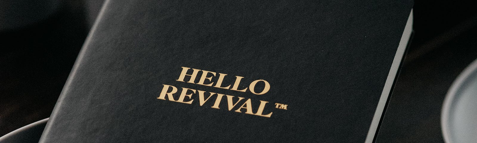A black book titled “Hello Revival” sitting in a white ceramic bowl surrounded by white ceramic mugs and plates