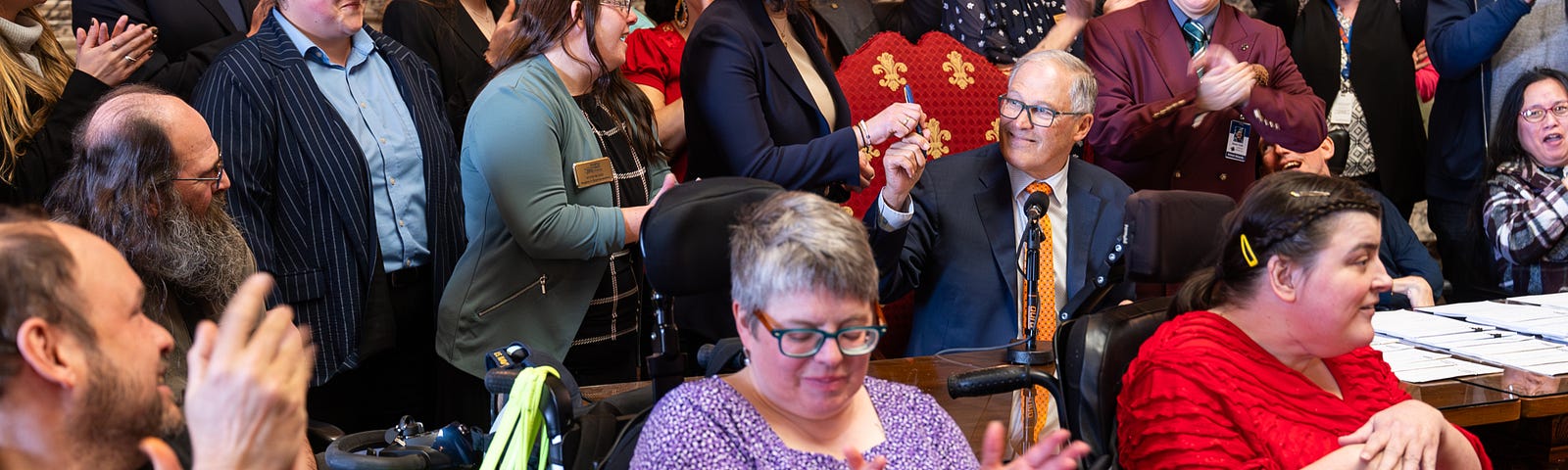 The image shows a bill signing. A group of people surround the governor and the large table at which he sits. The governor is wearing a blue suit with an orange tie and he is sitting in a chair upholstered in crimson and gold fabric. He is handing a pen to the woman on his right. The crowd of people (some of whom are in wheelchairs) are clapping and celebrating.