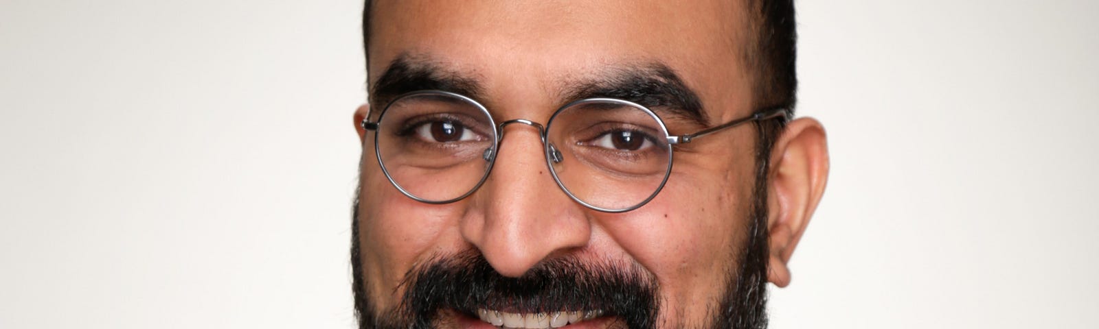 Headshot of a smiling man with black hair and a beard, and round glasses.