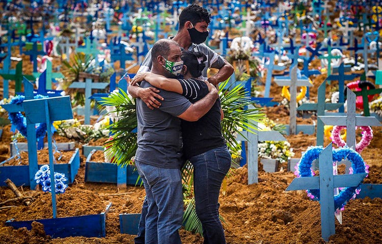 Na imagem há cruzes azuis e terra revolvida. Em destaque, três pessoas se abraçam enquanto choram. A foto é do cemitério de Manaus