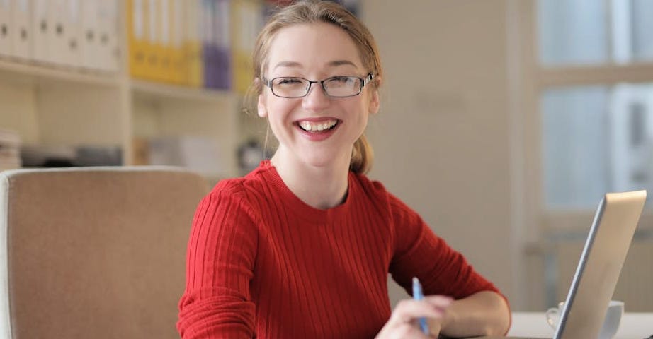 A woman wearing a burnt orange sweater and glasses at a computer with a pen in her hand.
