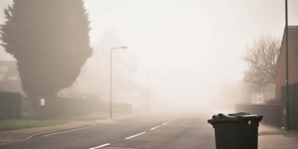 Trashcan on sidewalk next to a foggy residential street.