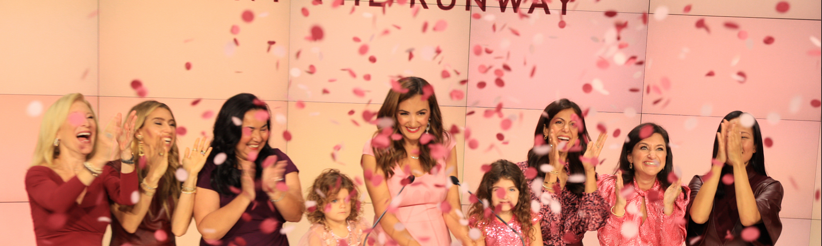 7 women and 2 girls cheering with pink confetti with “Rent the Runway” in background and foreground