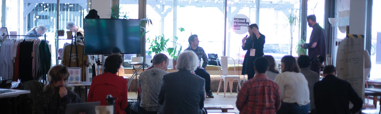 A speaker is standing and holding a microphone, whilst talking to another who is seated. They are in front of a large window that has a small round sticker that reads “Ethical Dilemma Cafe”. Clusters of audience members are seated on chairs and wooden benches in front of them.