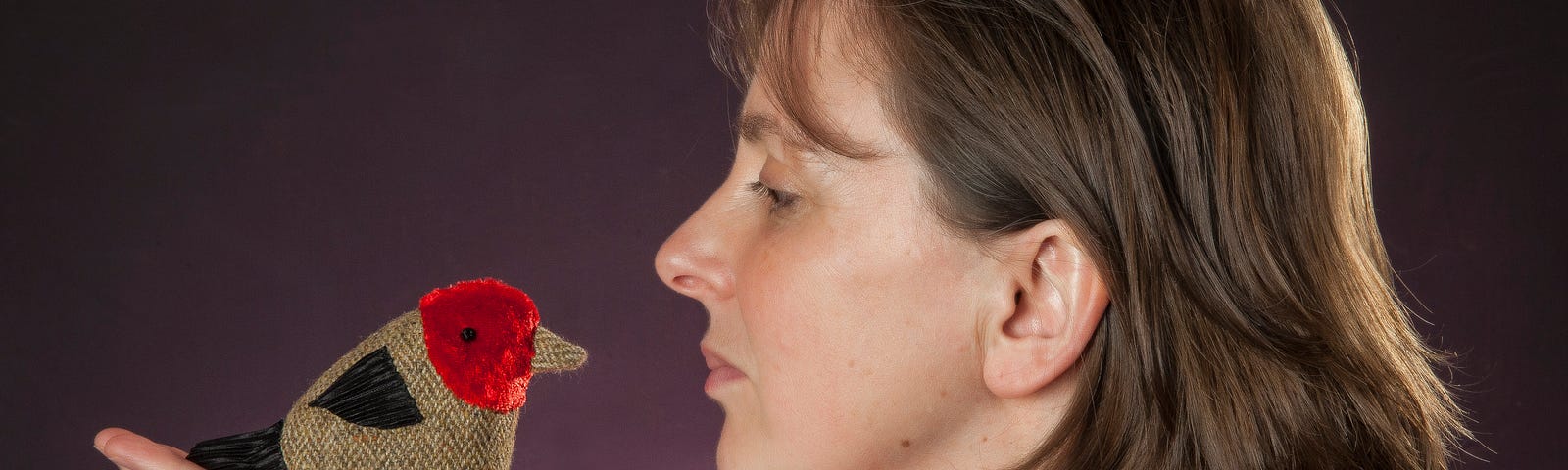 writer and storyteller Ailie Finlay looking at a fabric bird she holds in the palm of her hand, against a purple background