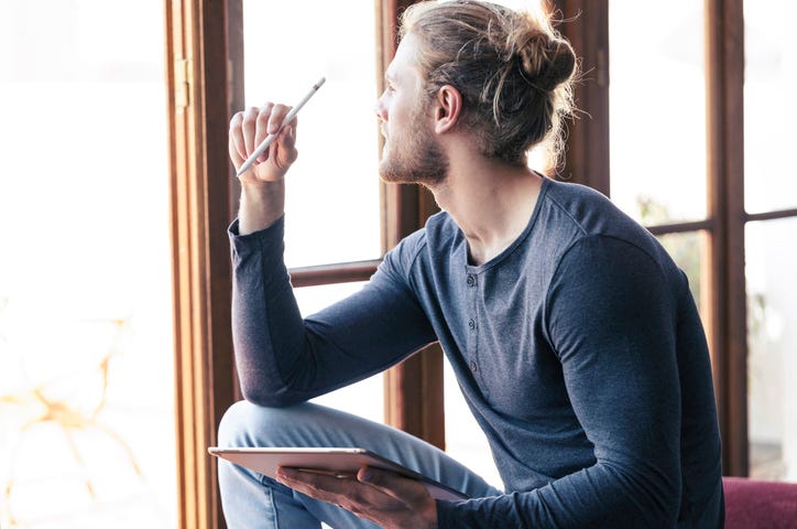Pensive young man at home with a digital tablet looking out of a window