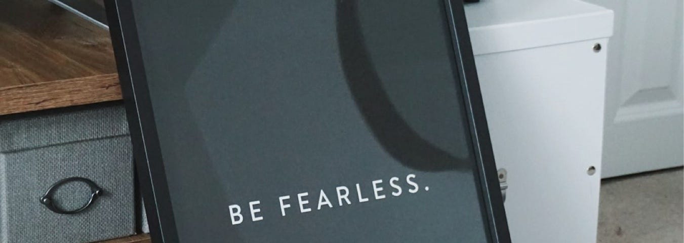 Leaning against a shelf, a black background framed in black shows just two words, written in capitals, a plain white font, that spells out “BE FEARLESS.”