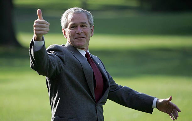 Former President George Bush Junior walking across the lawn in a suit, giving a thumbs up to the camera