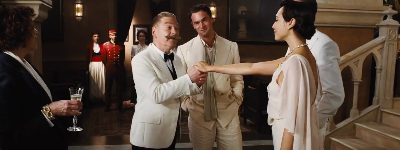 Kenneth Branagh (as Hercule Poirot) wearing a white tux and huge mustache takes Gal Gadot’s (Linnet Ridgeway)’s hand. She is wearing a white dress. Tom Bateman in a white suit is in the background between them, smiling.