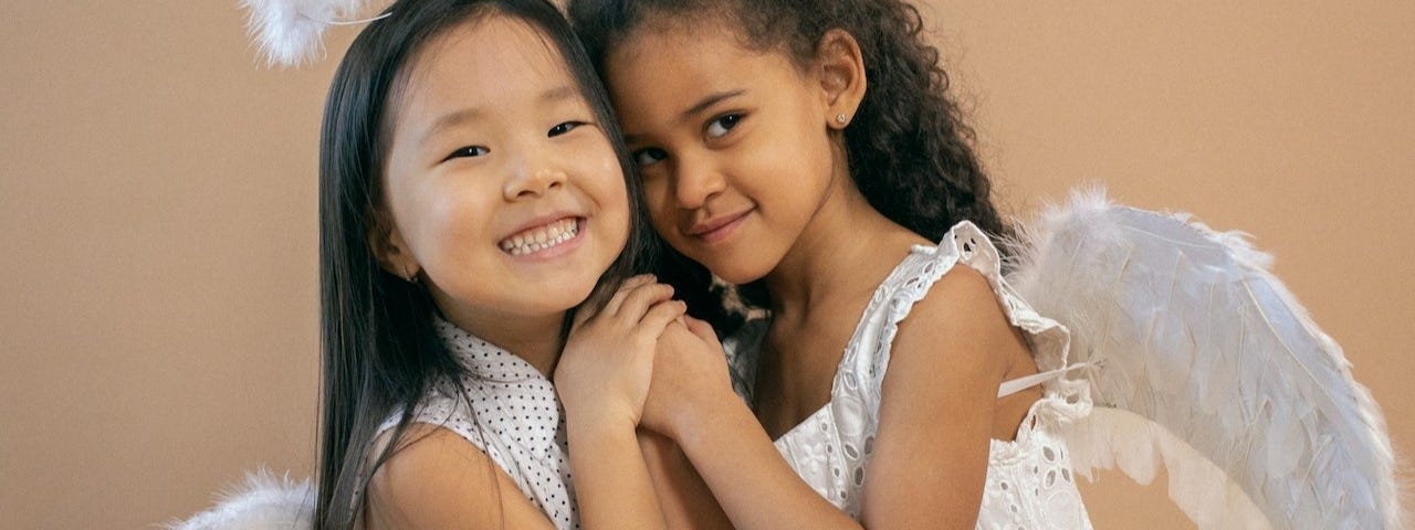 Two little girls, an Asian and a brown one, dressed in white as angels, hugging and smiling.