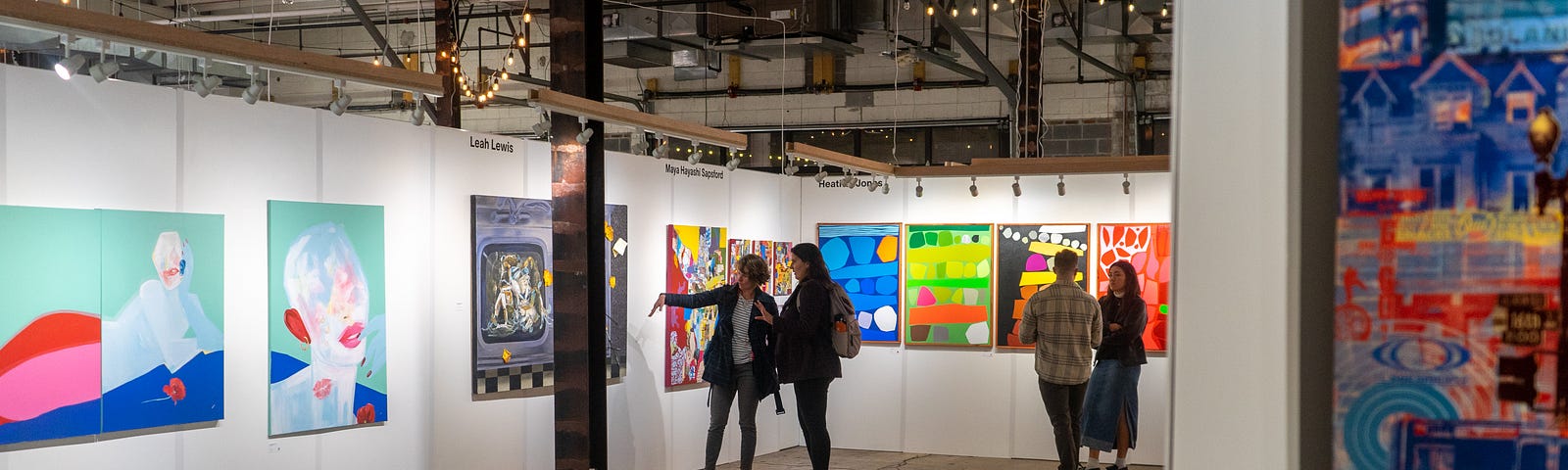 Colorful art hangs on walls inside an open warehouse-style space. Two groups stand and chat in front of the work.