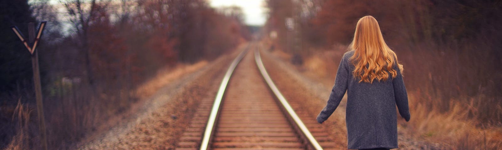 A woman with long red and a grey coat, balances on railroad tracks. Photo by Johannes Plenio on Unsplash