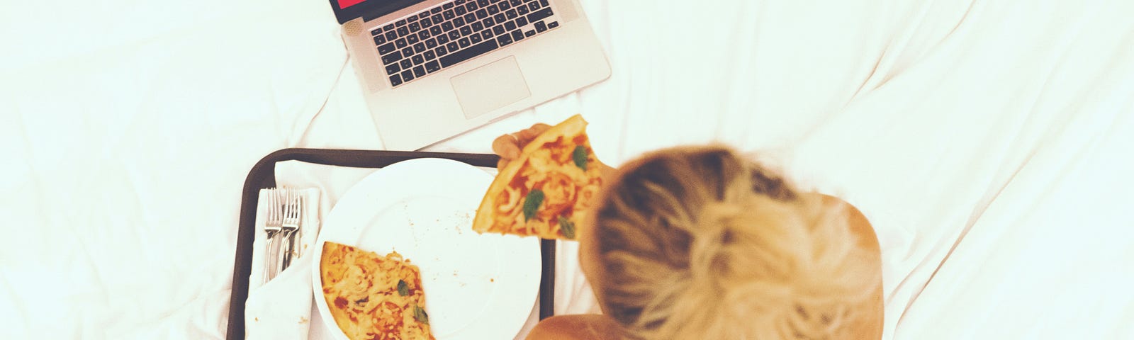 A lady sitting on a mattress, watching Netflix on a laptop and eating a slice of pizza.