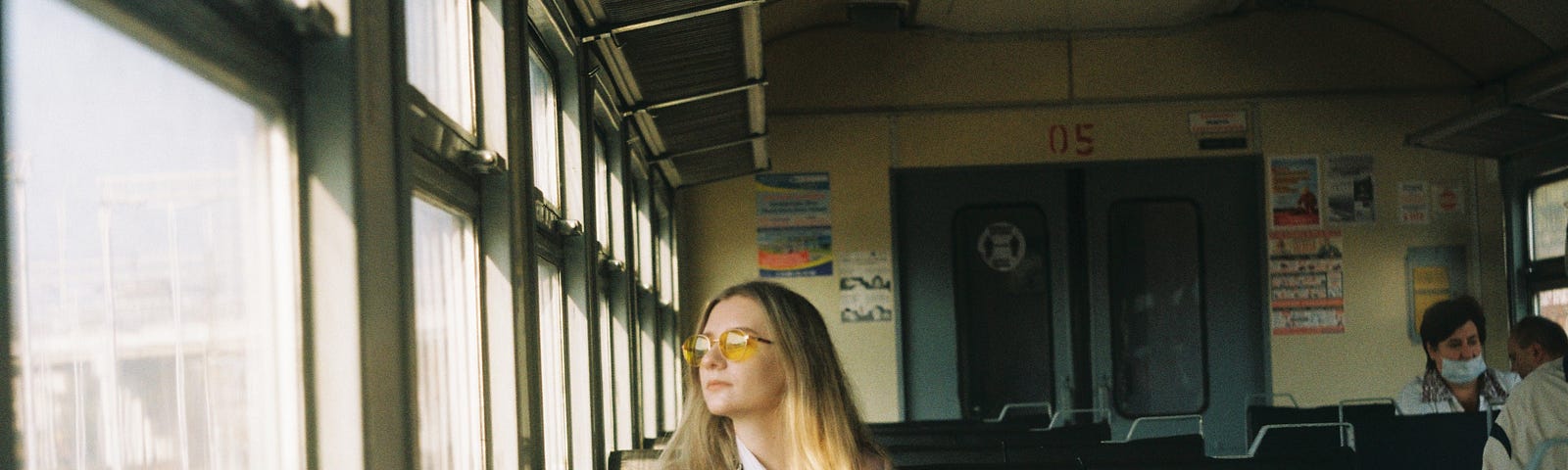 A lady in glasses, sitting in the window seat of a train, and sun is shining on her