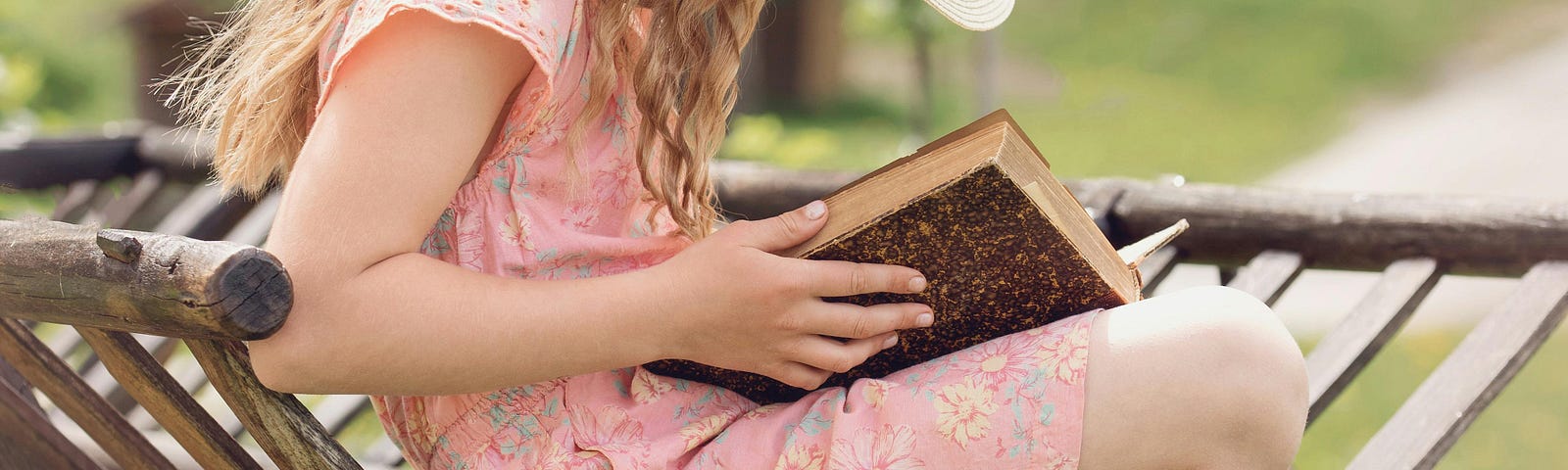 Image of a young girl wearing a pink dress and a white, floppy hat reading a book, sitting on a wooden wheelbarrow