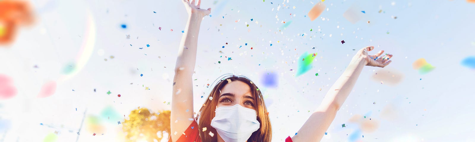 Woman wearing a face mask throws her arms up in celebration with her friends in the background and confetti in the air.