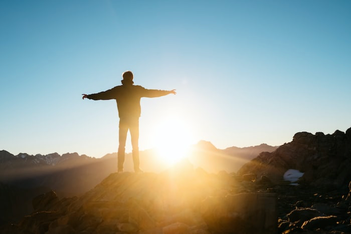 Photo of mountain climber on summit of a peak during the sunrise.