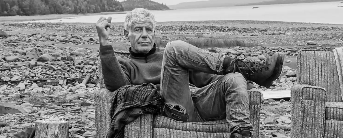 The image shows T.V chef, Anthony Bourdain sitting in an armchair on a rocky shore. He is dressed casually in a turtleneck, jeans, and boots, with one leg crossed over the other and a contemplative expression while holding a cigarette.