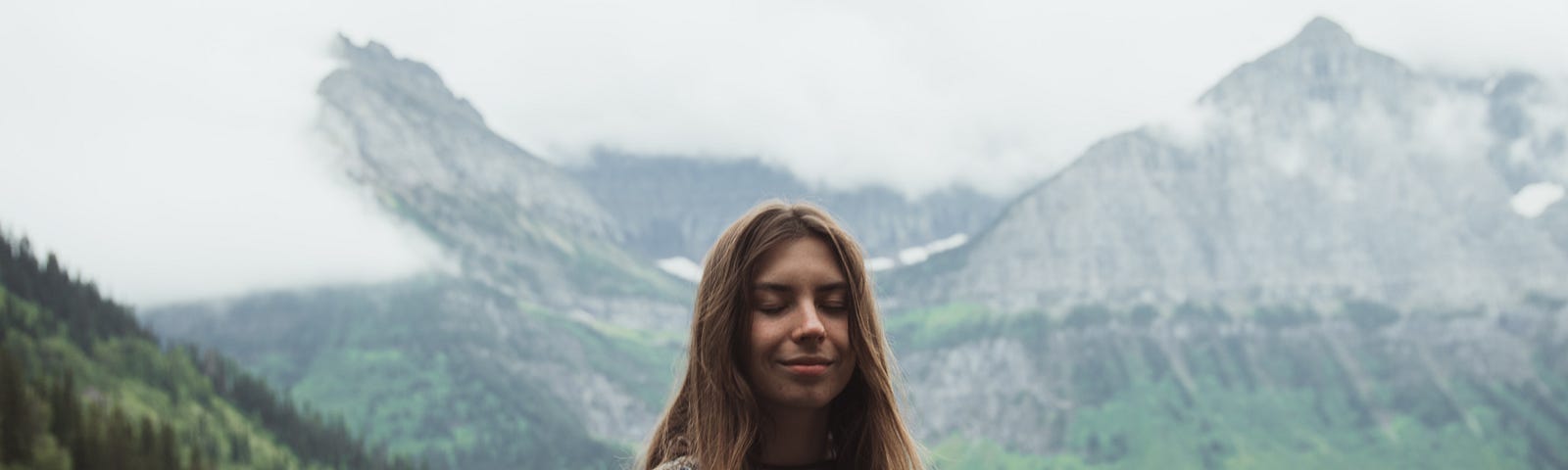 A woman meditates in a forest surrounded by mountains