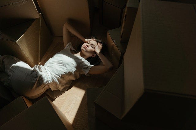 woman dressed in white lying amidst boxes, hands drawn up to her forehead. The Exhausting Adventure of Which Box to Choose is Emotional by Nancy Blackman. racial socialization, racism, other, othering, marginalization