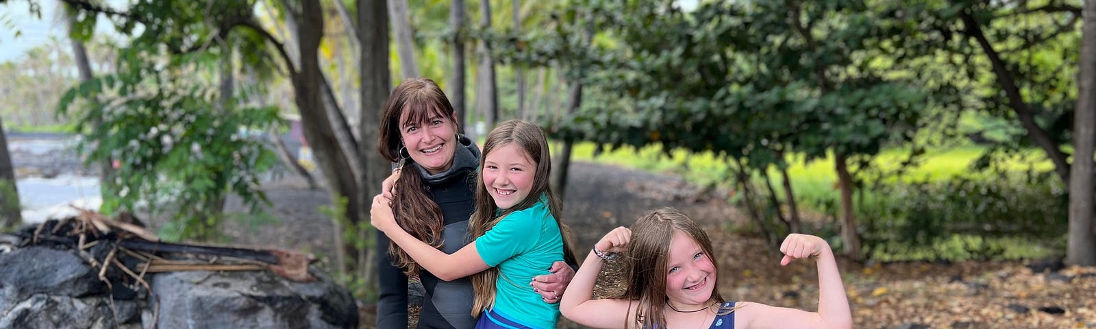 Mom in a wet suit holding a 10 year old girl in shorts and a younger sister showing her muscles. The beach is behind them and they’re being goofy.