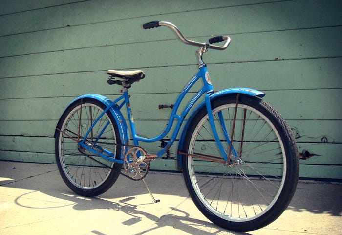 Vintage blue Schwinn girl’s bike in front of a wall with green siding.