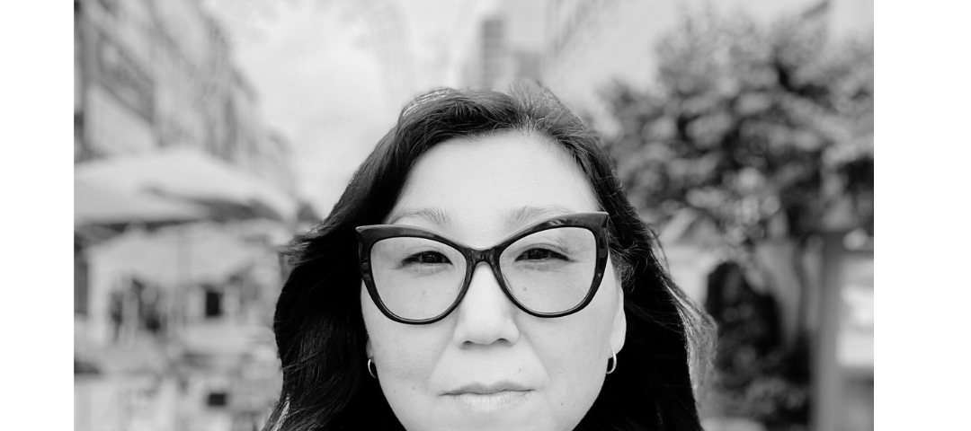 Black and white image of a Korean woman with large framed black butterfly glasses, long black hair oval face, in front of a blurry market.