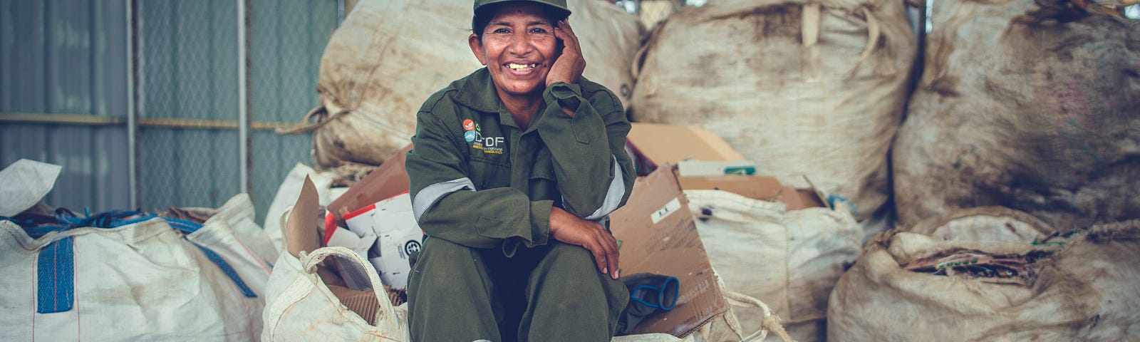 A woman dressed in work clothes smiles while sitting among neat piles, bags, and bundles of recycled items her company has collected.