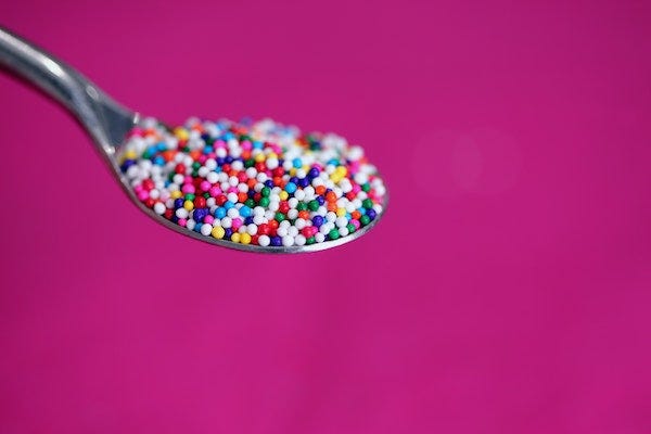 a teaspoon filled with rainbow-colored round sprinkles against a magenta background