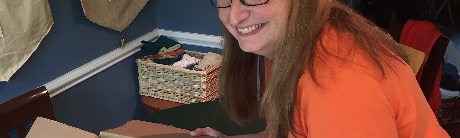 A smiling woman in an orange blouse displays a box of brightly colored books.