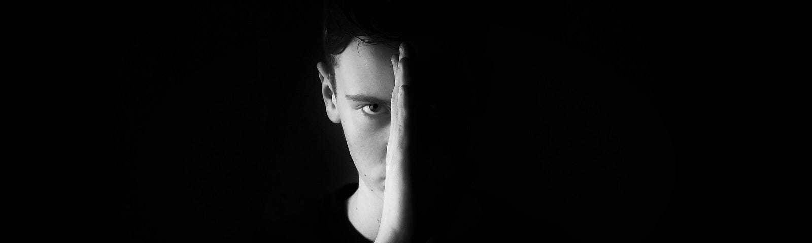 Whitesplaining goes on hiatus. A black and white photo of a young, dark-haired, white man facing forward, holding his hand up which divides his face equally in half. One side is in fully lit while the other half cannot be seen at all because its in complete darkness.