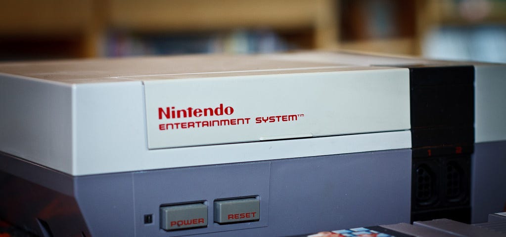 A close-up photo of a Nintendo Entertainment System console. A blurry Super Mario Bros. cartridge is in the bottom right, and some even blurrier cables (completely out of focus) are in the bottom left.