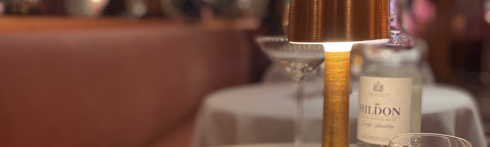 A table covered with a white mantel, in the center a gold lamp, a small pot with sugar sachets and a personalized hand sanitizer. a glass full of sparkling water to the right and to the left a black and gold menu.