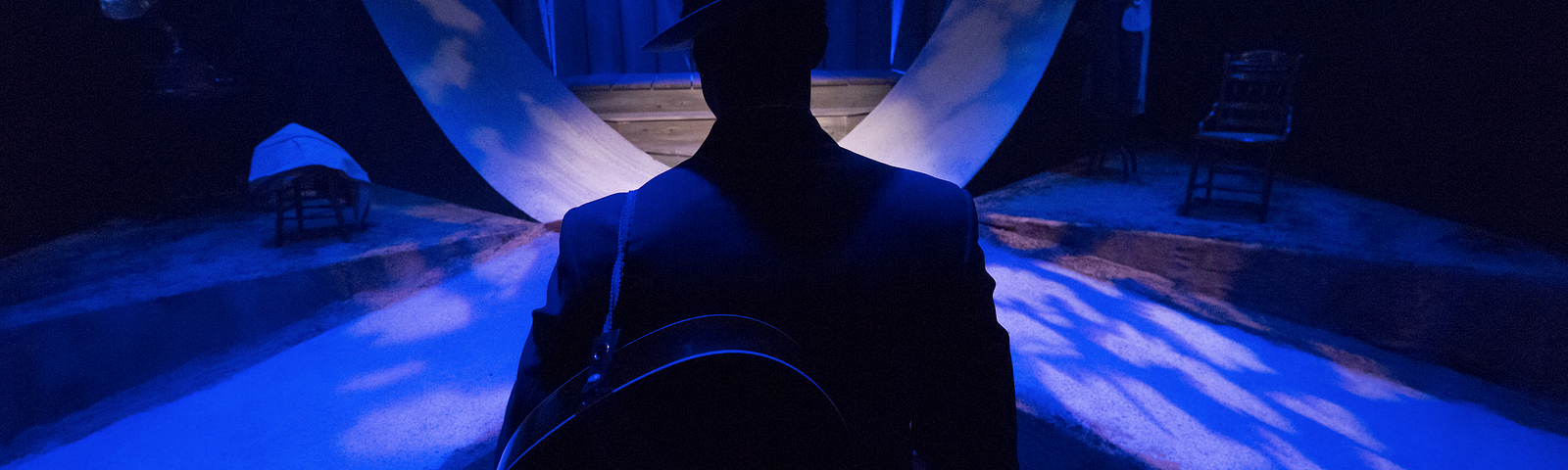 A man in a suit and hat with a guitar slung over his back stands with his back to the camera facing a blue-lit set